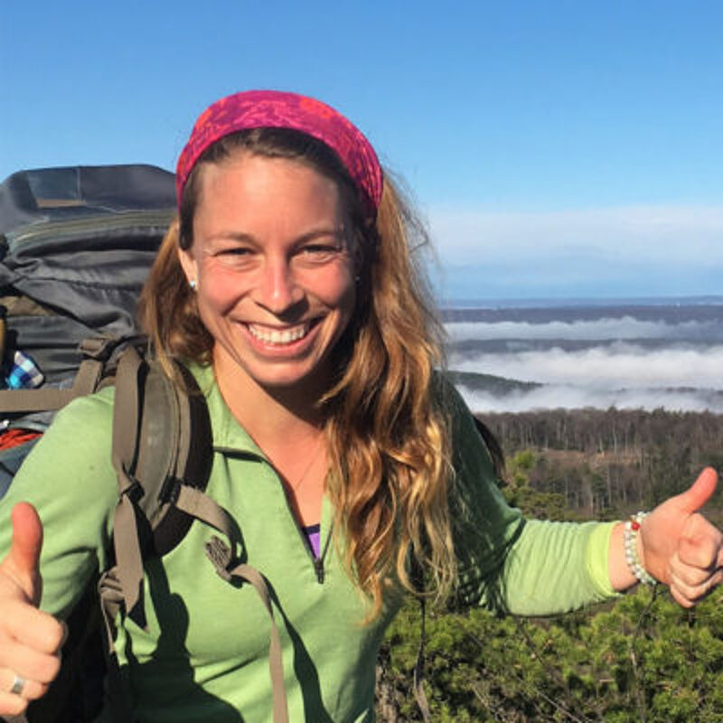 The image shows a woman with a backpack, giving a thumbs-up gesture. She is wearing a green jacket and a pink headband. The background features a scenic view with trees and a misty atmosphere. She appears to be enjoying the outdoors.